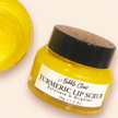 Glass clear jar with black lid of natural turmeric lip scrub revealing bright yellow hue of the lip scrub with partial image of open jar of the scrub highlighting soft creamy texture of the lip scrub. 