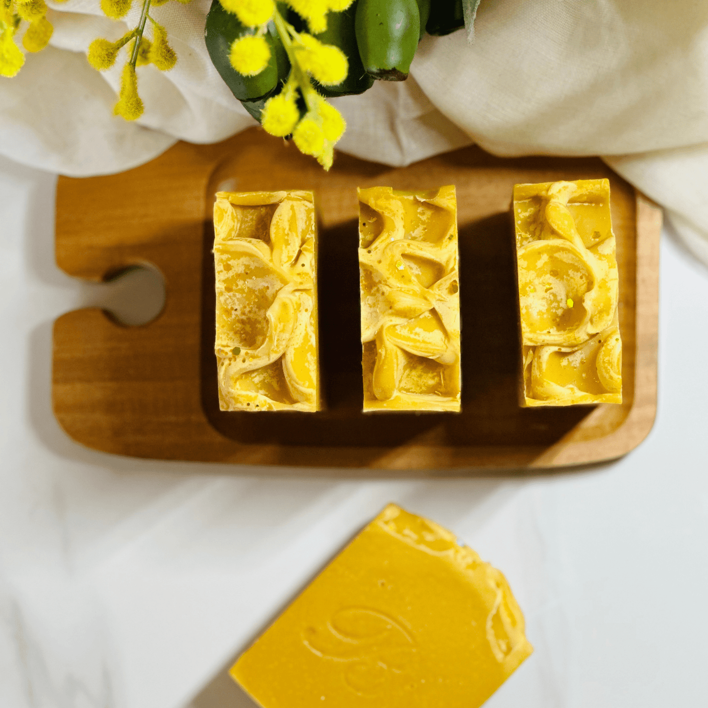 Bright yellow natural organic turmeric soap set on a wooden tray against a white background with botanicals and organic linen representing brightening organic  natural skincare.