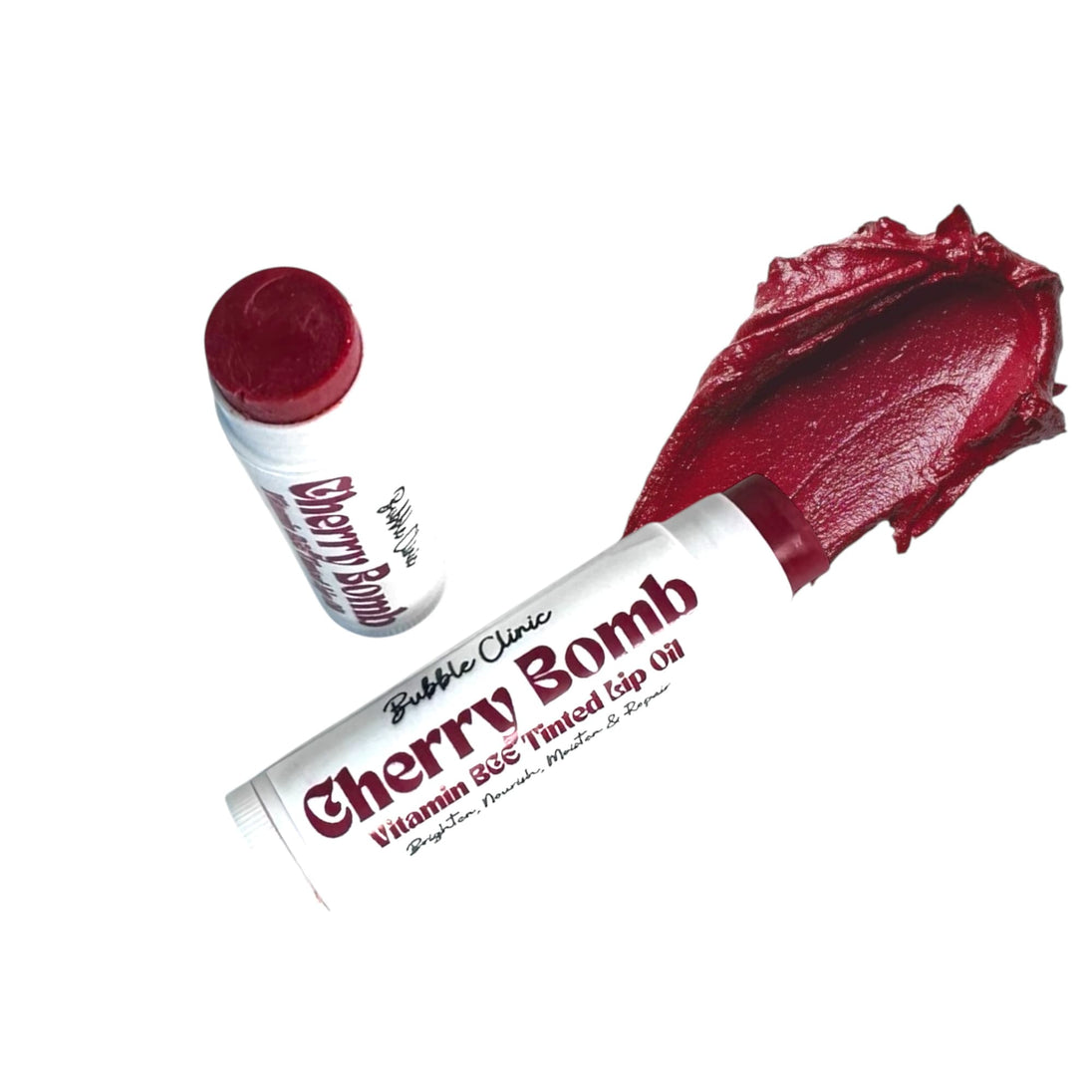 Open tubes of Cherry Bomb Tinted Lip Oil displayed against a clean white background, alongside a smooth, thick smear of the balm. The deep crimson shade highlights its rich pigment and silky, non-sticky texture.