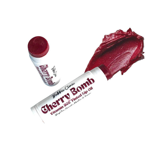 Open tubes of Cherry Bomb Tinted Lip Oil displayed against a clean white background, alongside a smooth, thick smear of the balm. The deep crimson shade highlights its rich pigment and silky, non-sticky texture.
