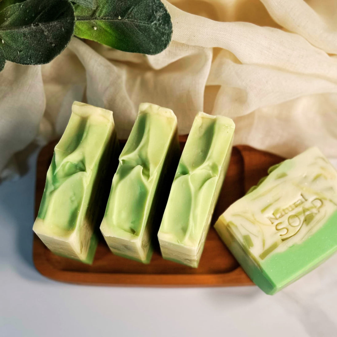 Four bars of brightening Kojic Acid & Alpha Arbutin Soap on an organic wooden tray, surrounded by lush green botanicals and soft natural cloth.