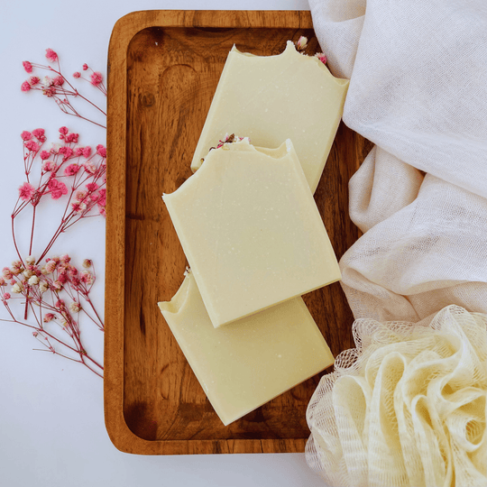 Front view of three natural goat milk soap bars on a wooden tray, surrounded by botanicals and an organic loofah, highlighting a natural skincare display.
