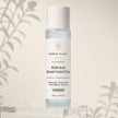 Bubble Clinic Advanced Multi-Acid Blemish Control Toner in a clear bottle with a white cap, displayed on a clear acrylic stand against a warm beige background with soft leaf shadows.