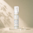 A bottle of Azeluxe Radiance Treatment byBubble Clinic on a beige surface with soft leaf shadows. The clear pump bottle is shown with its clear cap off, resting on a clear acrylic stand next to a small clear dish with the cream swatch on it.