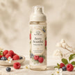 A bottle of Bondi Blooms 'Berry Beautiful' perfume body mist stands on a light stone surface, surrounded by fresh raspberries, blueberries, white blossoms, and soft beige leaves. The bottle has a cream-colored pump and a label decorated with illustrations of raspberries, blueberries, and green leaves.