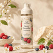 A bottle of Bondi Blooms Berry Beautiful Perfume Body Mist stands on a stone surface with raspberries, blueberries, white blossoms, and a woven textile in the background, creating a warm, natural aesthetic.