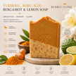 Two Tone Turmeric Kojic Acid brightening soap bar of Bubble Clinic Bergamot & Lemon Soap is displayed on a wooden stand, with a thick orange-brown top layer and a lighter beige lower layer. The background is a styled flat-lay featuring lemons, turmeric root, and green leaves, with text detailing the ingredients and product benefits.