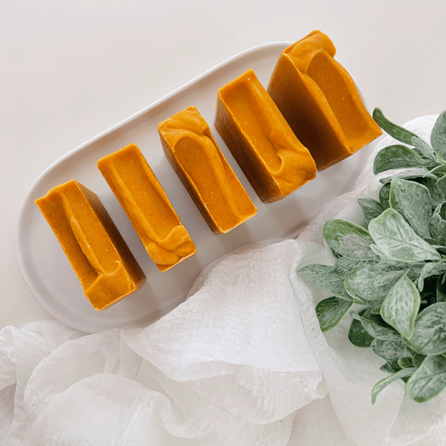Close-up of top of carrot turmeric kojic acid soap bars, highlighting their organic, clean texture, placed on a white concrete tray with organic leaves and light linen, representing clean beauty and natural skincare.