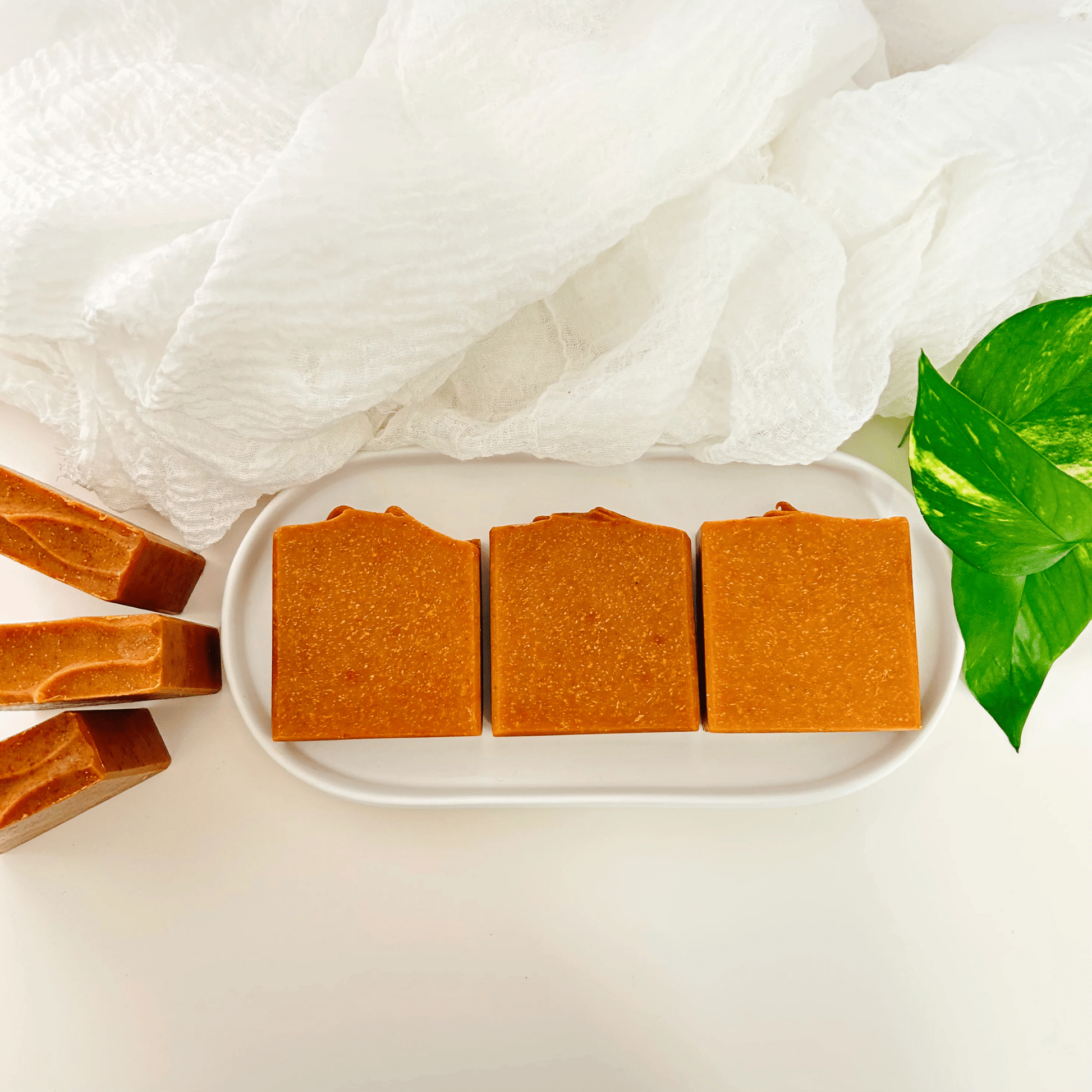 Carrot turmeric kojic acid soap bars laid flat, showcasing their bright, smooth, organic texture on a white concrete tray with organic leaves and light linen, symbolizing clean, natural skincare.