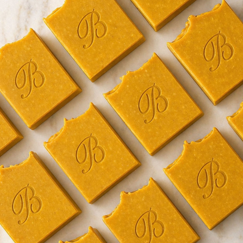 Multiple turmeric and kojic acid soap, flat laid and each embossed with a cursive letter B, arranged in a repeating pattern on a light marble-like surface.