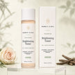 Bubble Clinic Advanced Brightening Toner bottle and matching outer box displayed on a clear acrylic stand, with a soft beige background and decorative rose and branch props.
