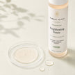 Bubble Clinic Brightening Toner bottle with a clear glass dish of pale cream serum and a few liquid droplets on a light beige surface with soft leaf shadows.