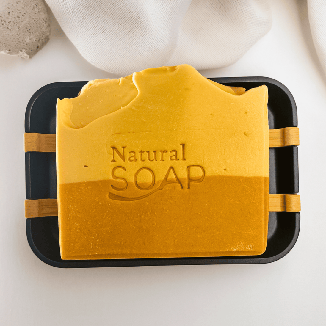 two tone brightening kojic acid soap with chamomile essential oil and turmeric on black soap dish against white background representing natural skincare.