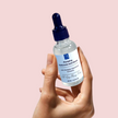 close up hand holding up glass serum bottle of brightening alpha arbutin serum with niacinamide against light pink background representing clean beauty rituals and natural australian brightening skincare
