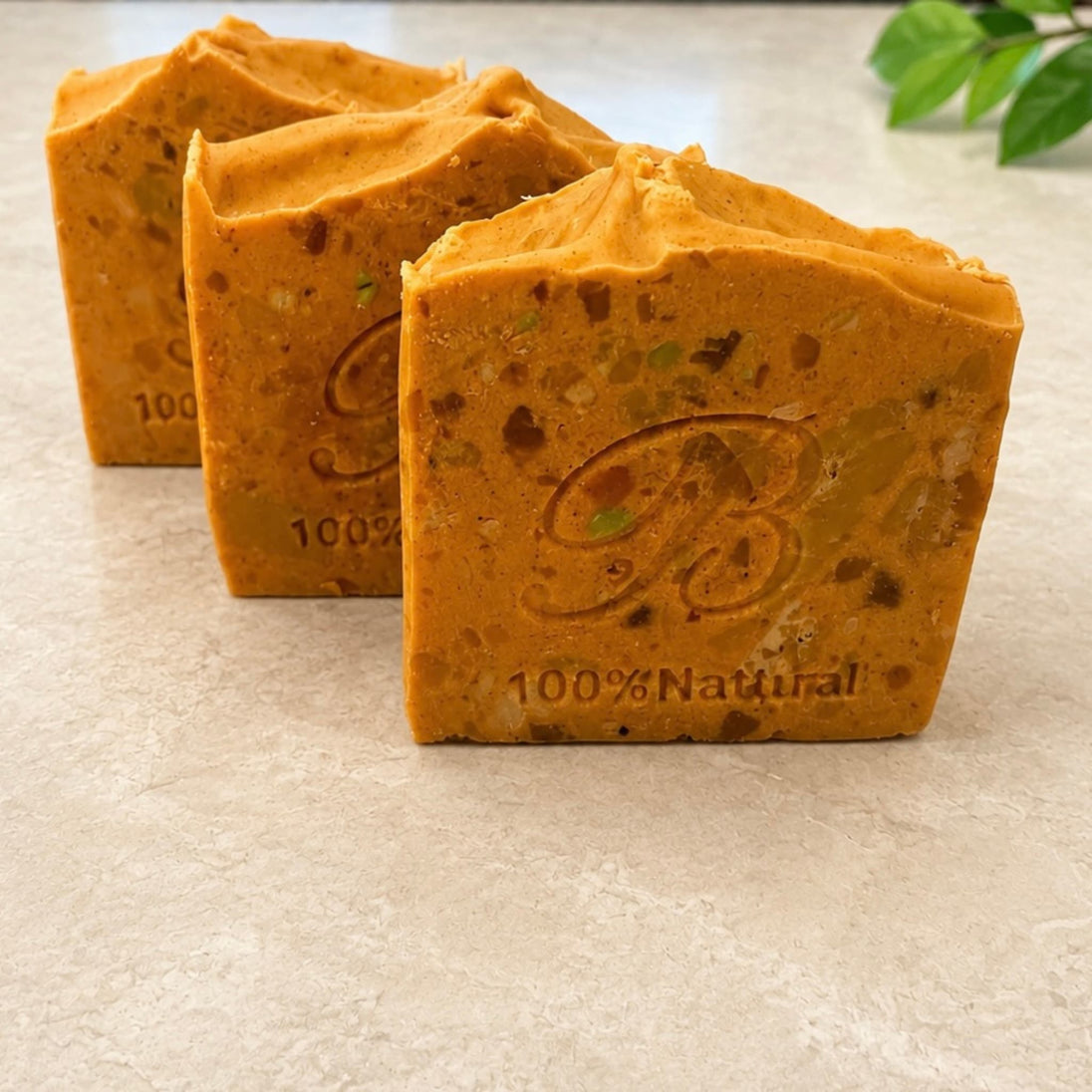 Three rectangular bars of handmade Turmeric soap with visible botanical inclusions, arranged on a light beige stone surface. The front bar is embossed with a large stylized 'B' and the text '100% Natural'.