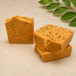 Three square bars of handmade turmeric soap with visible botanical inclusions, arranged on a light beige textured surface with a green leafy sprig in the background. One bar is embossed with a large 'B' and '100% Natural'.