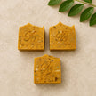 Three rectangular bars of handmade turmeric soap with kojic acid and alpha arbutin in a warm golden-yellow color, arranged on a light beige textured surface with green leaves in the top right corner. Each bar is embossed with a monogram logo and the text '100% Natural'.