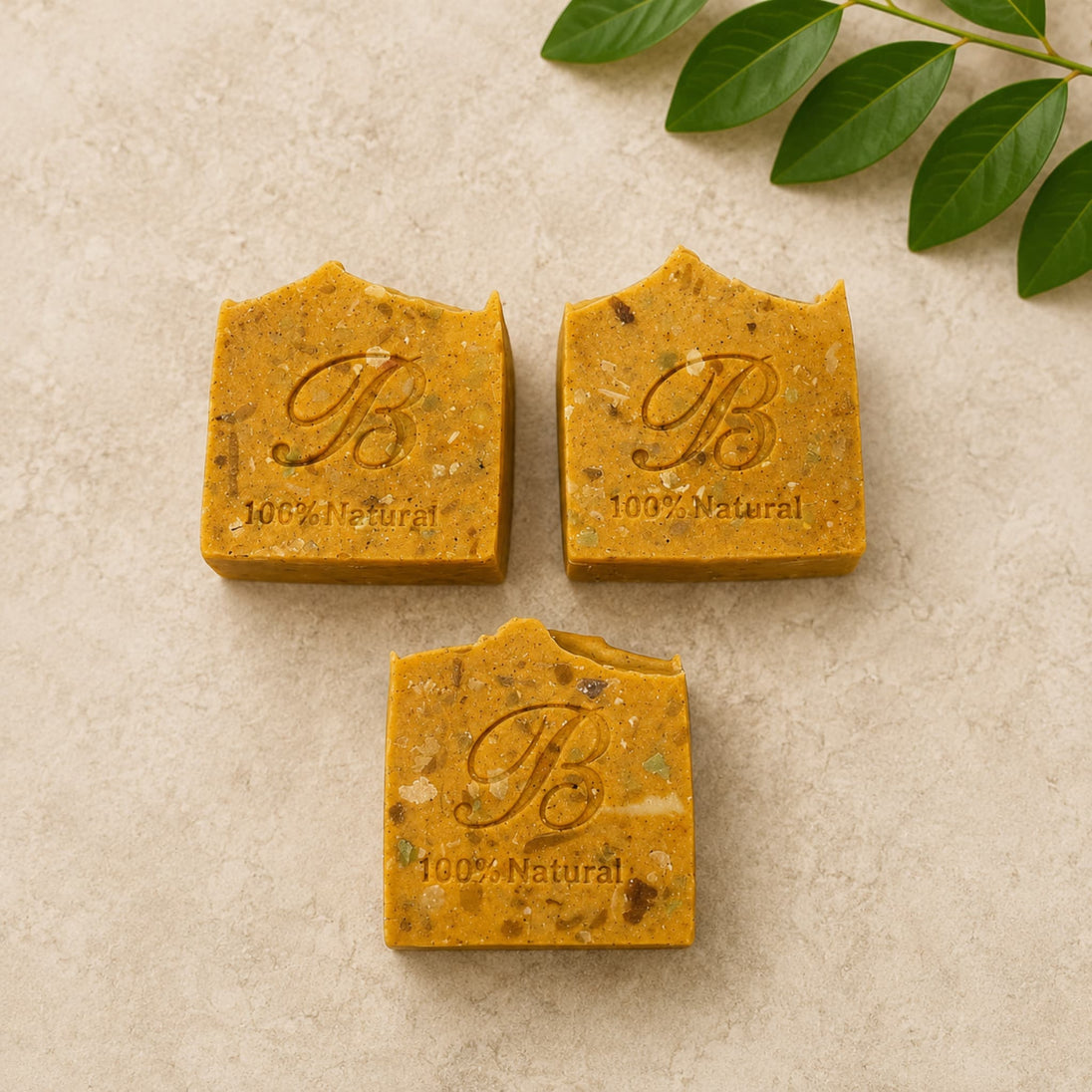 Three rectangular bars of handmade turmeric soap with kojic acid and alpha arbutin in a warm golden-yellow color, arranged on a light beige textured surface with green leaves in the top right corner. Each bar is embossed with a monogram logo and the text '100% Natural'.