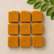 Nine square kojic and turmeric soap bars  arranged in a 3x3 grid on a light beige textured surface, with green leaves in the top-right corner. Each bar is embossed with a large script letter 'B' and the words '100% Natural'.