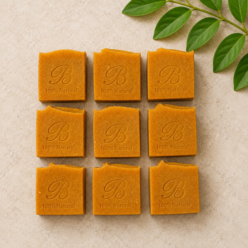 Nine square kojic and turmeric soap bars  arranged in a 3x3 grid on a light beige textured surface, with green leaves in the top-right corner. Each bar is embossed with a large script letter 'B' and the words '100% Natural'.