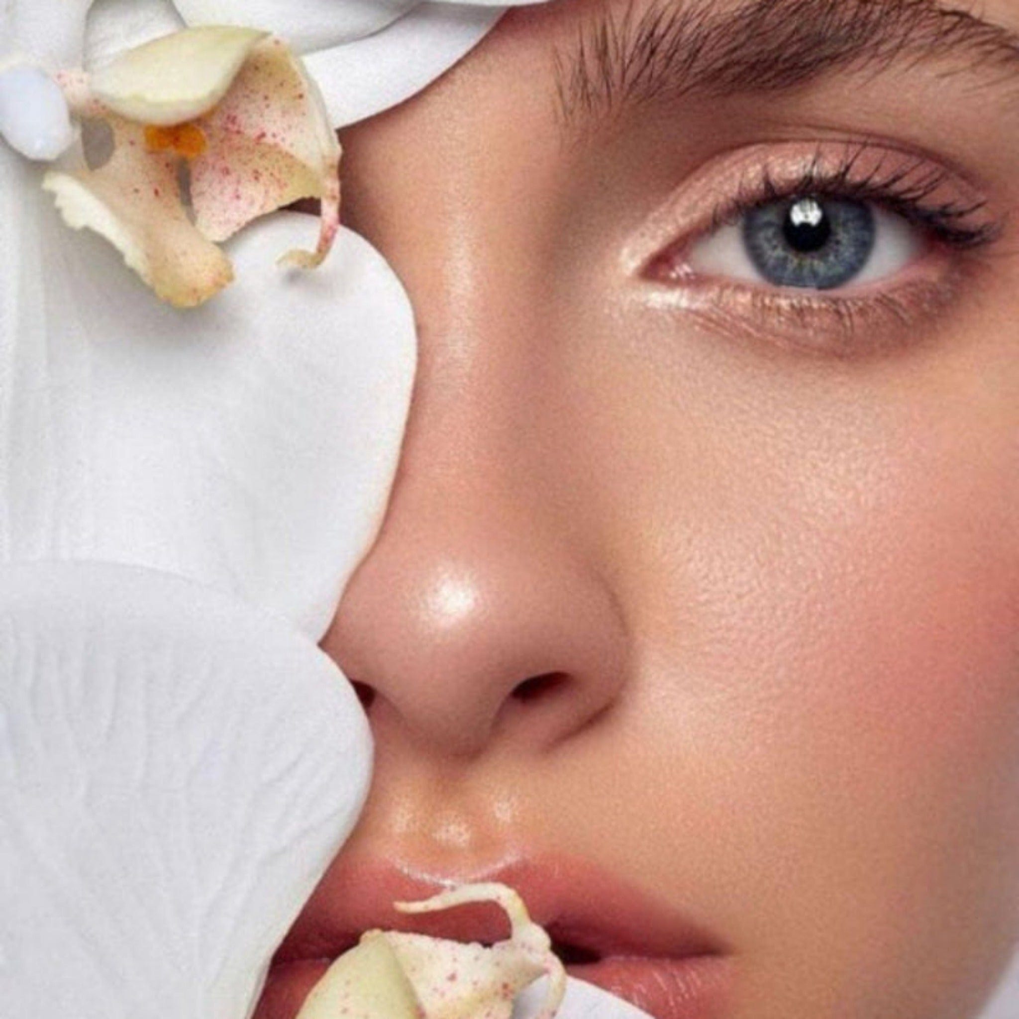 Close-up beauty shot of a woman's face with a white orchid flower resting against her cheek, highlighting smooth skin, blue eye, and soft pink lips.