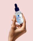 close up of hand holding up luxury glass bottle of plumping brightening alpha arbutin serum with niacinamide against pink background representing natural australian skincare for radiant skin