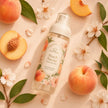 A clear fine mist body perfume bottle labeled 'Peach Nectar' is arranged on a peach-toned surface with whole and sliced peaches, green leaves, and white blossoms.