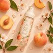 A clear fine mist body perfume bottle labeled 'Peach Nectar' is arranged on a peach-toned surface with whole and sliced peaches, green leaves, and white blossoms.
