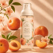 A bottle of Peach Nectar Perfume Body Mist stands on a stone surface surrounded by fresh peaches, peach slices, and blossoms. The bottle has a white cap and a cream label decorated with peach illustrations.