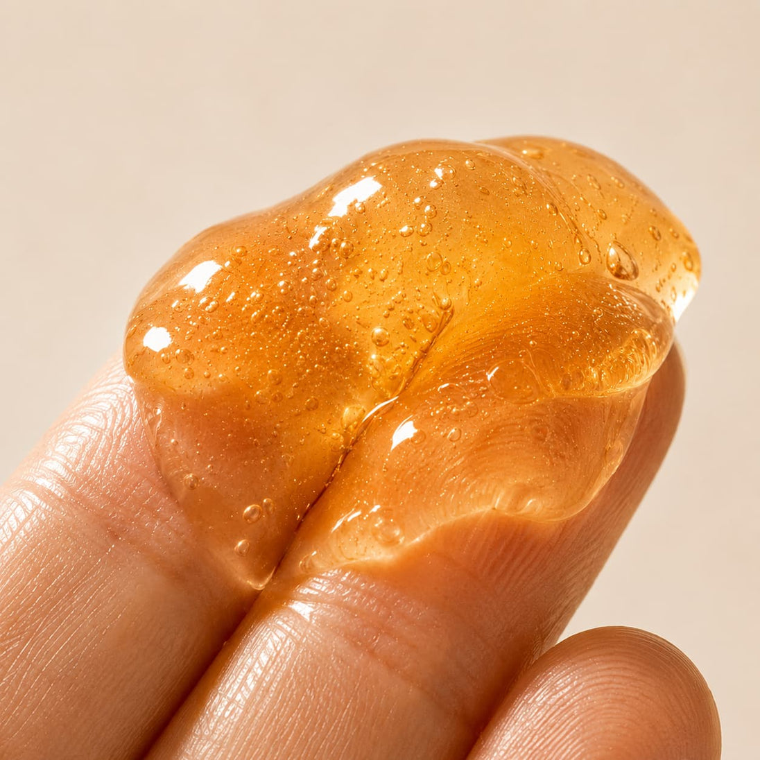 Close-up macro photograph of the Bubble Clinic Peptides Anti Aging Jelly Mask texture resting between fingertips, showcasing the translucent amber jelly with suspended hydration bubbles and a rich, glossy gel consistency under soft studio lighting.