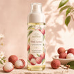 A bottle of Bondi Blooms Pink Lychee Perfume Body Mist stands on a stone surface with lychee fruits and leaves arranged around it. The bottle has a white cap and a cream label decorated with pink lychee illustrations, set against a warm beige background with soft leaf shadows.