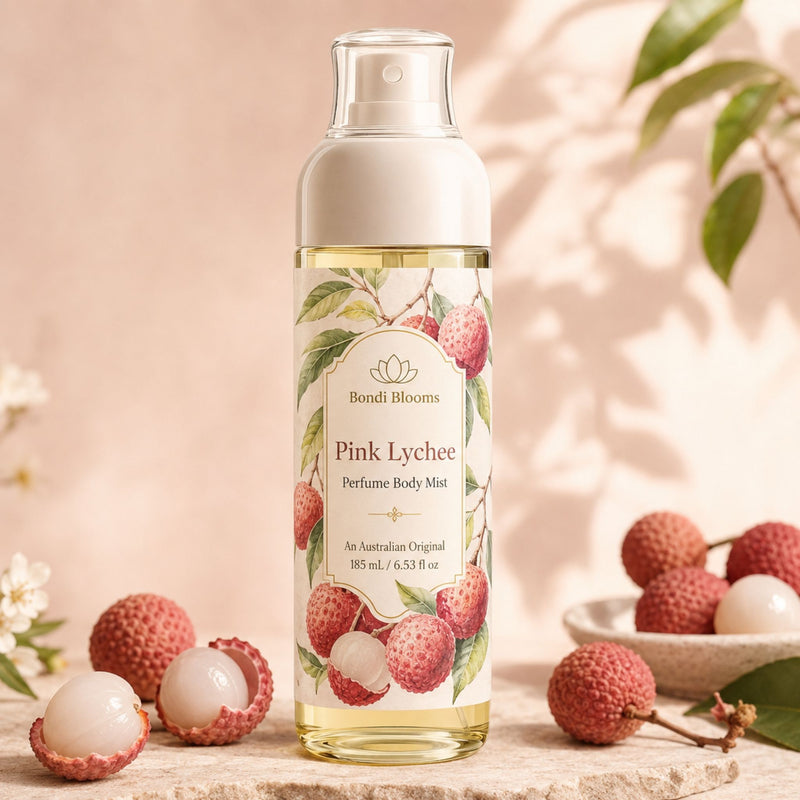 A bottle of Bondi Blooms Pink Lychee Perfume Body Mist stands on a stone surface with lychee fruits and leaves arranged around it. The bottle has a white cap and a cream label decorated with pink lychee illustrations, set against a warm beige background with soft leaf shadows.