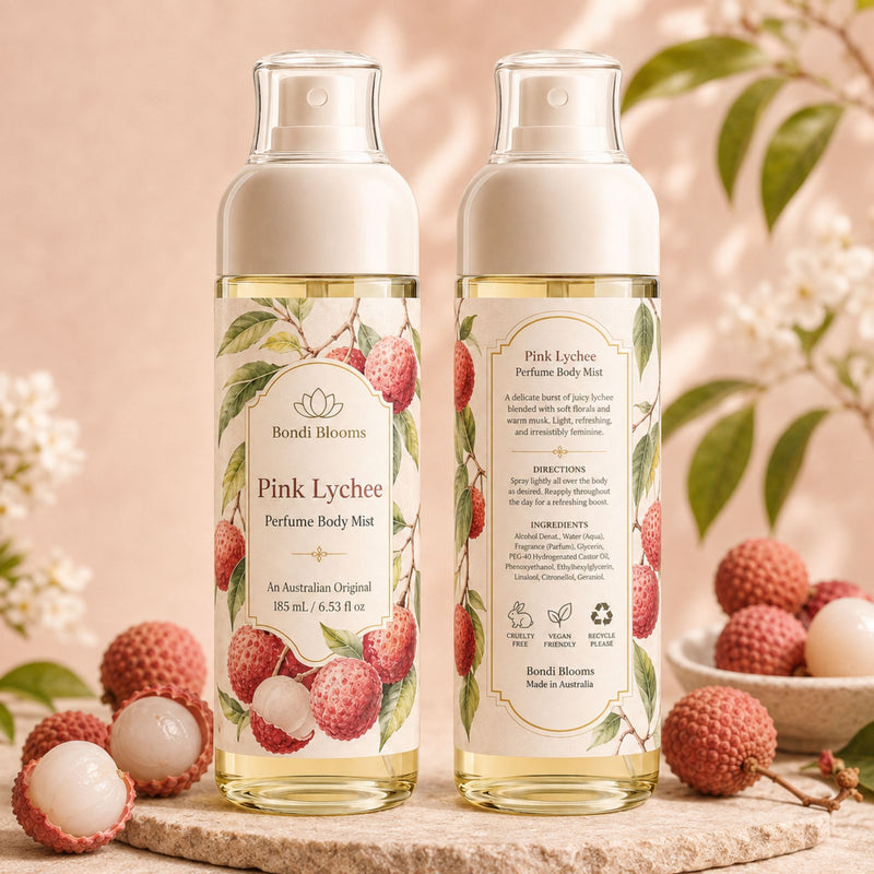 Two bottles of Bondi Blooms Pink Lychee Perfume Body Mist shown on a stone surface with lychee fruits and soft floral background props. The bottles are clear with pale yellow liquid inside, white caps, and labels featuring pink lychee illustrations.