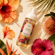 A bottle of Samba Perfume Body Mist by styled in a flat-lay with tropical hibiscus flowers and palm leaves on a warm beige background.