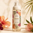 A bottle of Samba Perfume Body Mist stands centered on a warm beige surface with tropical flowers and leaves arranged around it. The bottle is clear glass with a white pump cap and a colorful floral label featuring orange, pink, green, and cream tones.