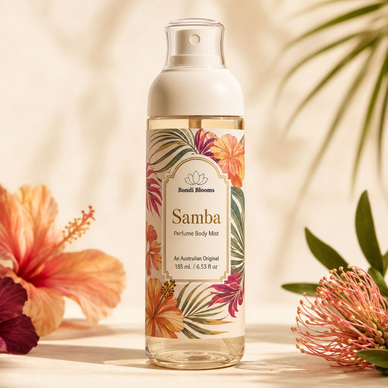 A bottle of Samba Perfume Body Mist stands centered on a warm beige surface with tropical flowers and leaves arranged around it. The bottle is clear glass with a white pump cap and a colorful floral label featuring orange, pink, green, and cream tones.