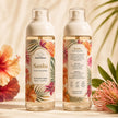 Two clear fine mist bottles of Bondi Blooms Samba Perfume Body Mist, showing both the front and back labels, set against a warm beige background with tropical flowers and leaves.