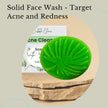 Green solid face wash bar on a stone surface with text about targeting acne and redness.