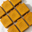 Several rectangular bars of handmade turmeric and kojic soap in a warm golden-brown tone, arranged diagonally on a white marble surface. Each bar is embossed with a decorative monogram letter 'B' representing best turmeric soap australia bubble clinic. 