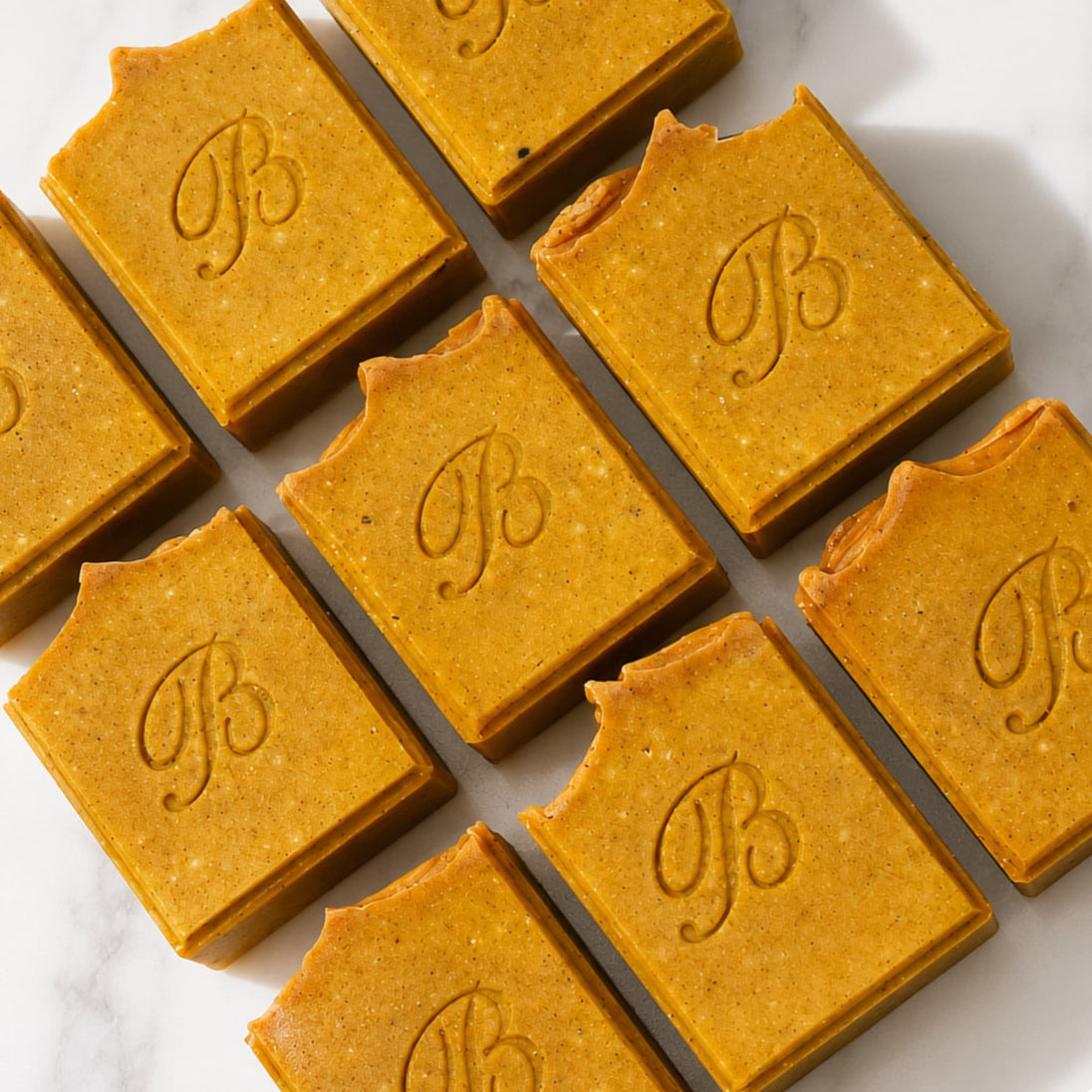 Several rectangular bars of handmade turmeric and kojic soap in a warm golden-brown tone, arranged diagonally on a white marble surface. Each bar is embossed with a decorative monogram letter 'B' representing best turmeric soap australia bubble clinic. 