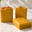 Three textured handmade turmeric and kojic soap bars in a warm golden-brown color, featuring an embossed monogram 'B' on the front, arranged on a light marble surface with soft leaf shadows representing best brightening turmeric soap australia.