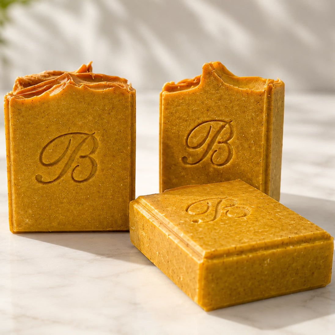Three textured handmade turmeric and kojic soap bars in a warm golden-brown color, featuring an embossed monogram 'B' on the front, arranged on a light marble surface with soft leaf shadows representing best brightening turmeric soap australia.