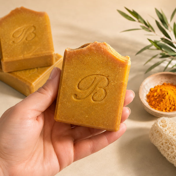 A hand holds a square bar of golden-turmeic kojic soap embossed with a script letter 'B'. More soap bars and a small bowl of turmeric powder sit on a beige surface with a sponge and green leaves in the background representing best turmeric kojic soap australia.