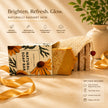 A boxed natural turmeric soap bar set displayed on a warm beige surface with decorative ribbons and a vase in the background. The set includes a floral soap bar in a clear container wrapped with burlap, a matching branded box, and a gift information panel.