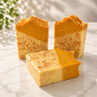 Three artisanal handcrafted turmeric soap soap bars with a two-tone design of orange and pale yellow, featuring a speckled pattern and an embossed monogram 'B', displayed on a light marble surface representing best turmeric brightening soap australia.