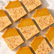 Several rectangular bars of handmade turmeric and kojic acid soap arranged on a light marble surface. Each bar has a warm orange-brown top half and a pale cream lower half with scattered pinkish speckles, and a decorative monogram 'B' embossed on the front.
