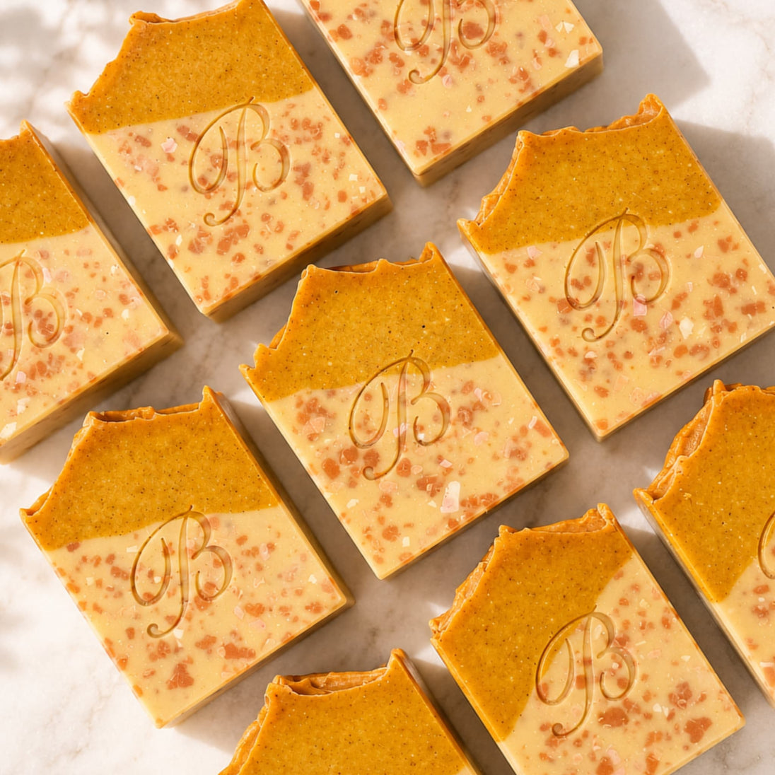 Several rectangular bars of handmade turmeric and kojic acid soap arranged on a light marble surface. Each bar has a warm orange-brown top half and a pale cream lower half with scattered pinkish speckles, and a decorative monogram 'B' embossed on the front.
