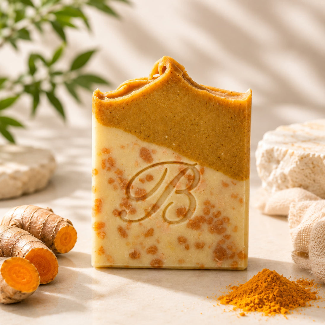 Close up of a rectangular handmade turmeric kojic acid soap bar with a wavy top edge, featuring a two-tone design of golden-brown and creamy beige with orange speckles. The letter 'B' is embossed on the front.