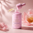 Pink bottle of Luminous Vitamin C Brightening Serum Intense with a pink dropper cap and glass dropper, styled with pink flowers on a soft pink background.